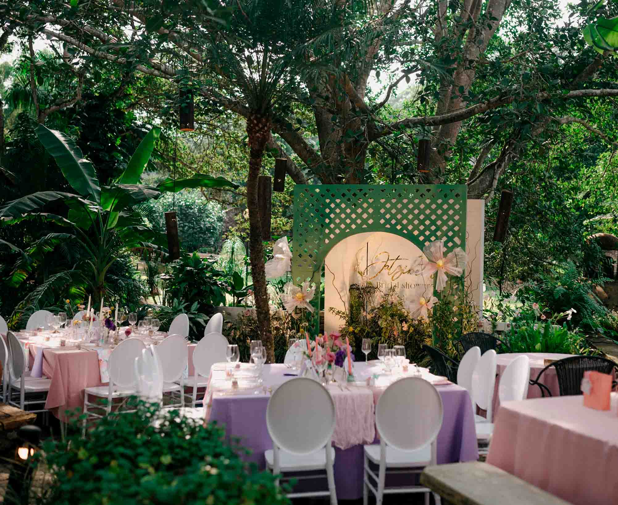 Jardín para eventos en Quinta Pichón, con mobiliario como mesas con manteles de tonos pasteles en rosa y purpura, el jardín esta rodeado de arboles y distintas especies de plantas de varios tamaños.