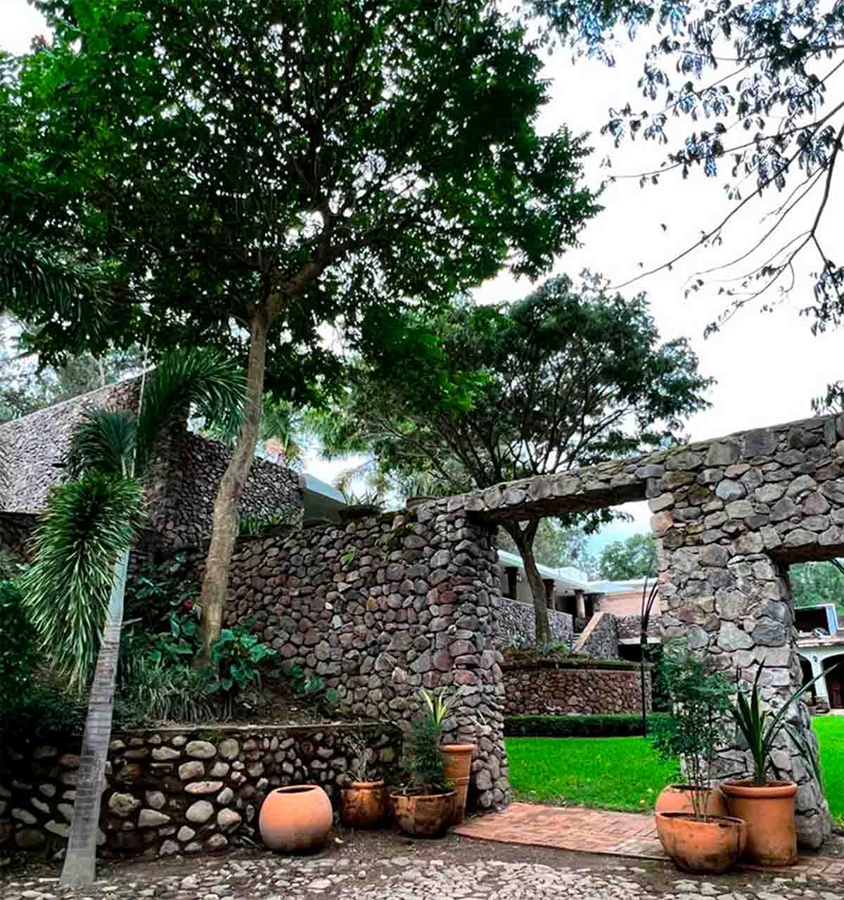 Entrada en forma de arco hecha en mampostería con árboles y macetas con plantas; la entrada lleva a la zona de habitaciones del Hotel Quinta Pichon.