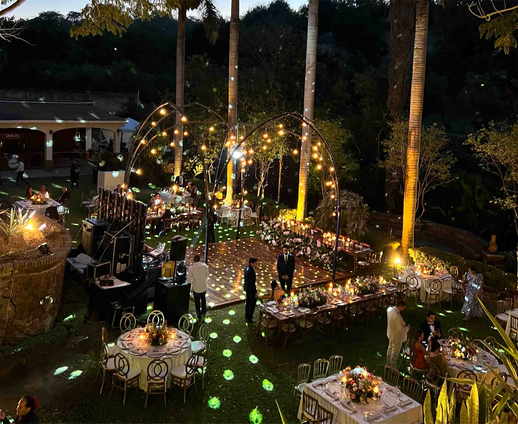 Jardín de Quinta Pichón con mobiliario como mesas con manteles blancos y sillas con respaldos circulares, el lugar esta decorado con luces al igual el arco metálico en el centro decorado con luces para algún evento social.