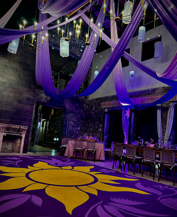 Instalaciones interiores de la casona, zona en Quinta Pichón utilizada para llevar acabo eventos sociales, decorada con adornos en tonos morados y en el centro un gran sol sobre el suelo, toda la sala tiene iluminación en tonos morados.