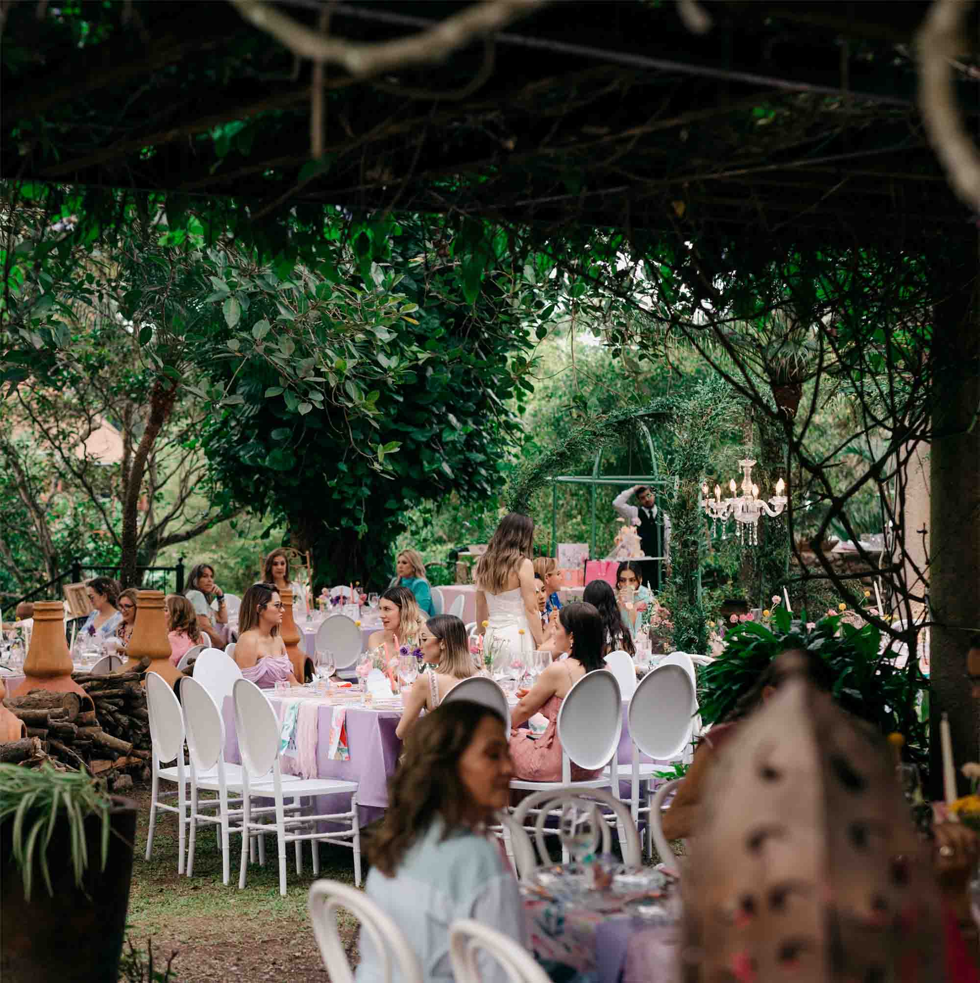 Jardín para eventos en Quinta Pichón, con mobiliario como mesas con manteles de tonos pasteles en rosa y purpura, el jardín esta rodeado de arboles y hay invitados presentes en su totalidad mujeres.