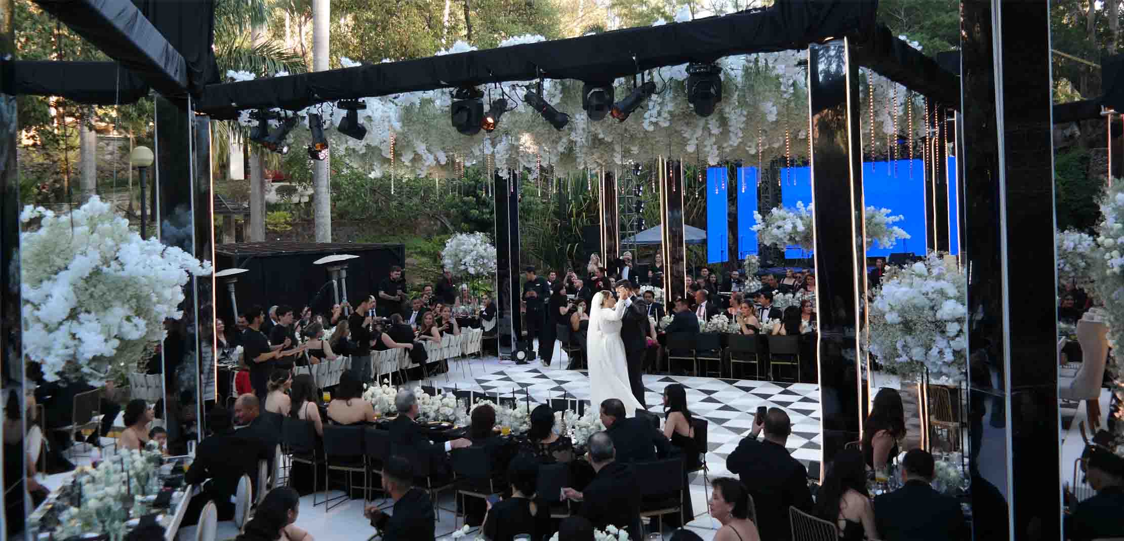 Boda celebrada en el jardín principal de Quinta Pichón, en el centro un gran escenario con una estructura color negro para sostener la iluminación, los invitados están situados alrededor del escenario principal donde la pareja de novios esta bailando.