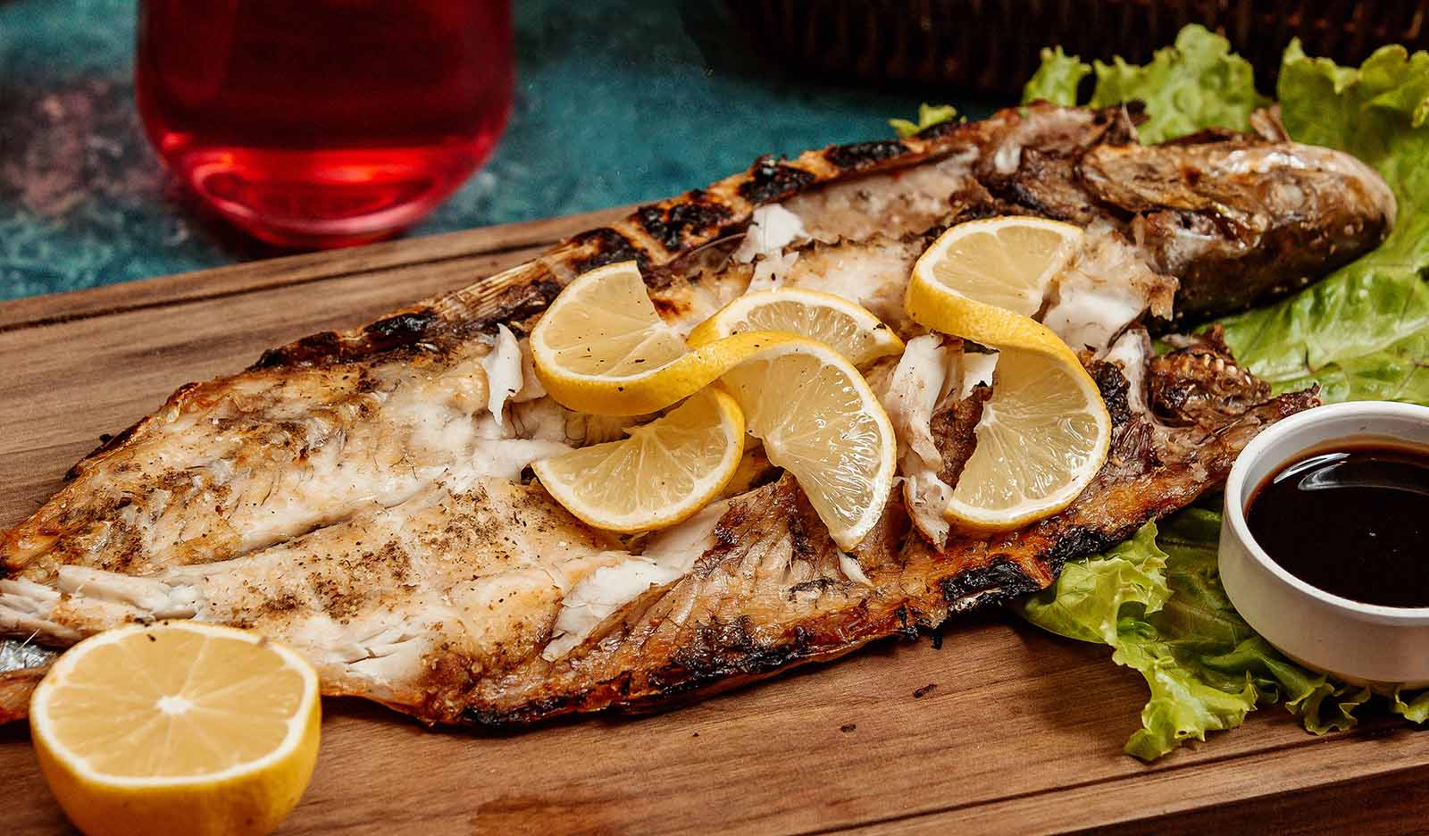 Pescado cocinado estilo a las brazas acompañado con rodajas de naranja y lechuga para decorar, este esta servido en un plato de madera.