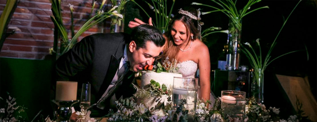Pareja de novios celebrando su boda, el novio esta apunto de darle una mordida al pastel y su novia trata de empujar su rostro hacia el pastel.