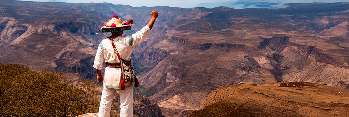 Un huichole de espaldas en la cima de una montaña viendo el paisaje.