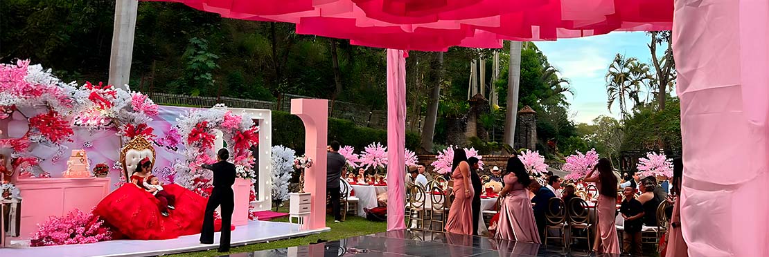 Mujer joven celebrando sus 15 años, viste un vestido rojo y el escenario esta adornado de con flores color blancas y rojas, hay adornos de color rosa que combinan con la demás decoración.