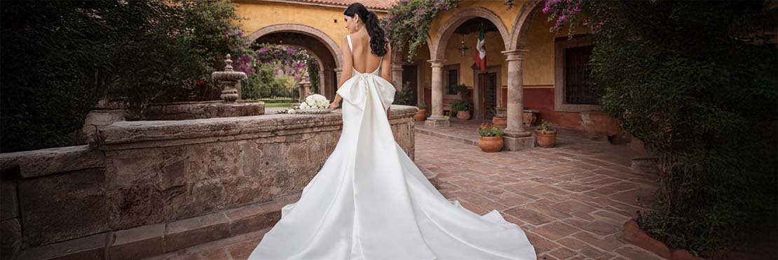 Novia posando para una fotografía viste un vestido blanco con un velo de color blanco, aun lado de ella sobre una fuente de piedra hay un ramo de flores color blanco.